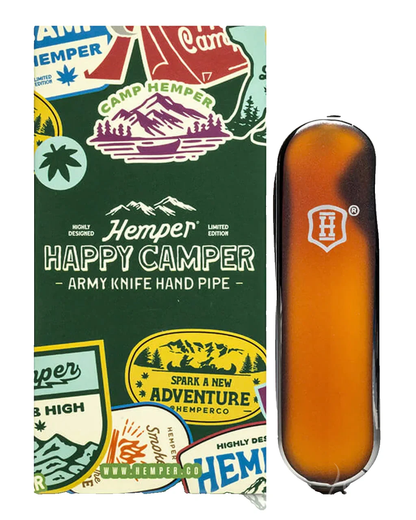 A Hemper Happy Camper Army Knife Hand Pipe shown next to its illustrated packaging box. The hand pipe is made of amber-colored glass with a dark metallic rim and a white shield logo.