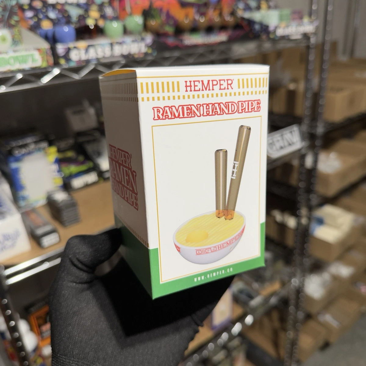 A hand wearing a black glove holds a miniature collectible box of 'Hemper Ramen Hand Pipe' figurines. The packaging features a retro design with yellow, white, and green sections, and an illustration of a bowl of ramen with two hand pipes.