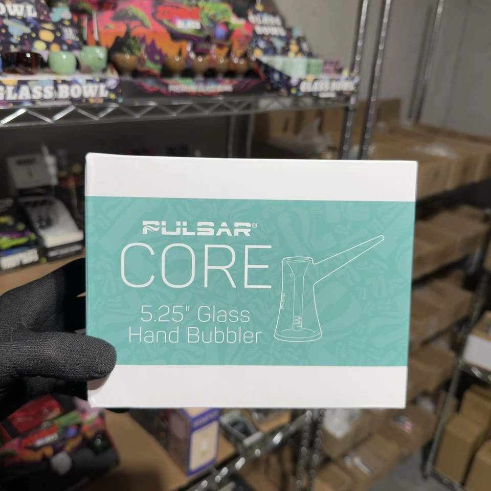 A hand wearing a black glove holds a white and teal Pulsar Core 5.25" Glass Hand Bubblower box in a cluttered warehouse or store setting with metal shelving and cardboard boxes in the background.